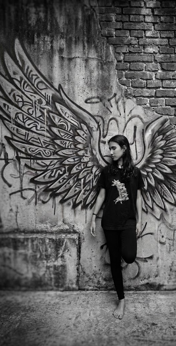 Helena Richter encostada em um muro com asas pintadas, fotografia em preto e branco